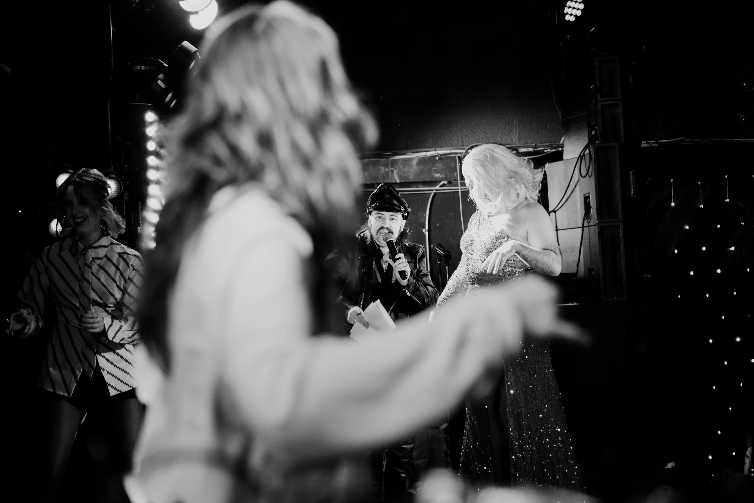 Black and white photo of a stage performance. One woman in a striped shirt and woman in a sparkly dress are visible, along with a man in a leather jacket and police hat holding a microphone, possibly singing or speaking. Audience members are in the foreground, slightly out of focus.