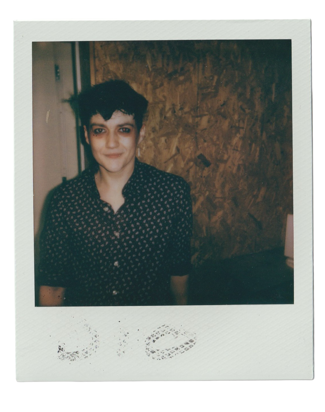A young woman with short black hair and tattoos on her right eyebrow and left cheek, standing indoors against a wooden wall, wearing a black polka dot shirt.