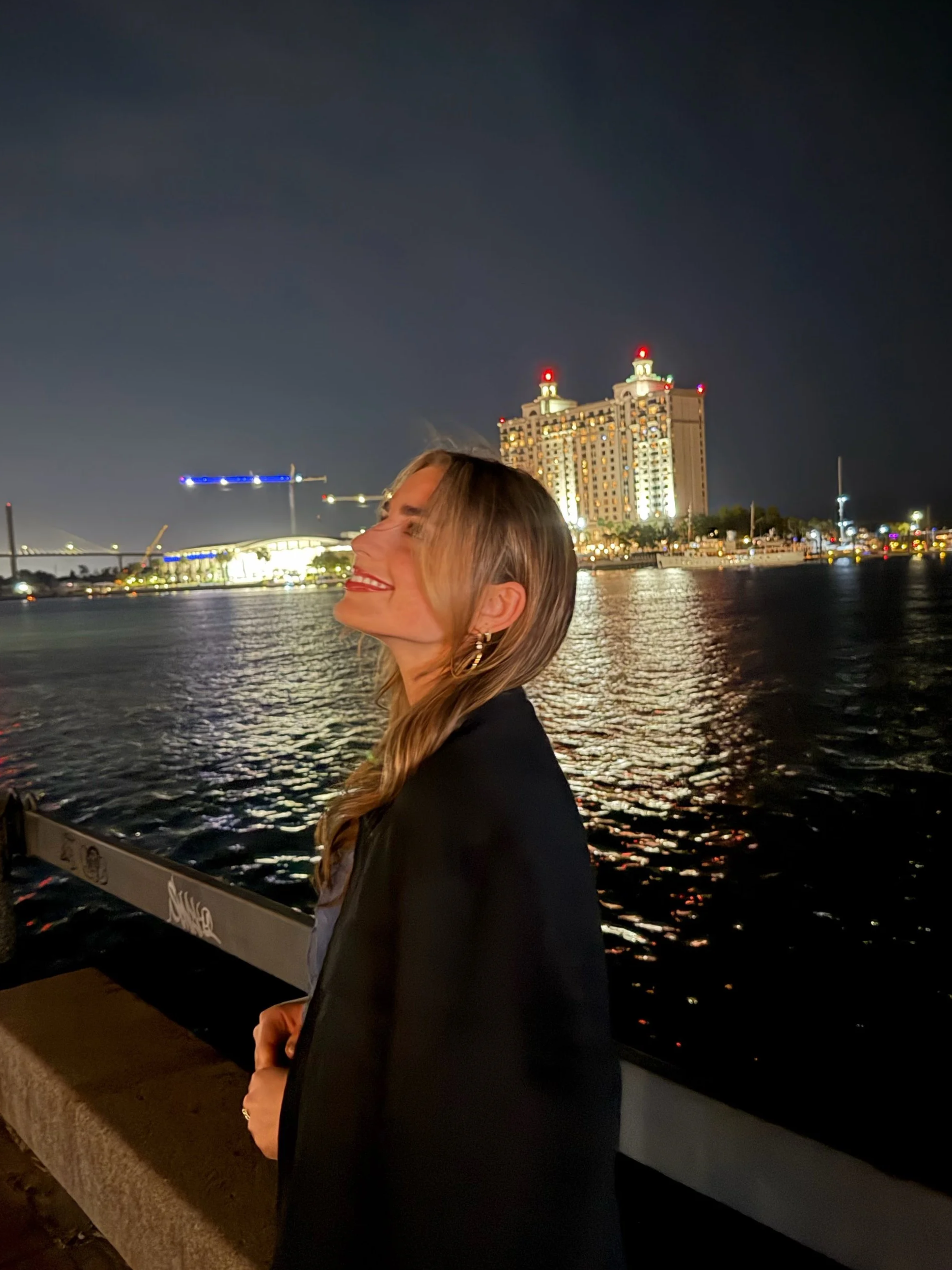 Woman smiling with her eyes closed, standing near water at night, with city lights and tall buildings in the background.