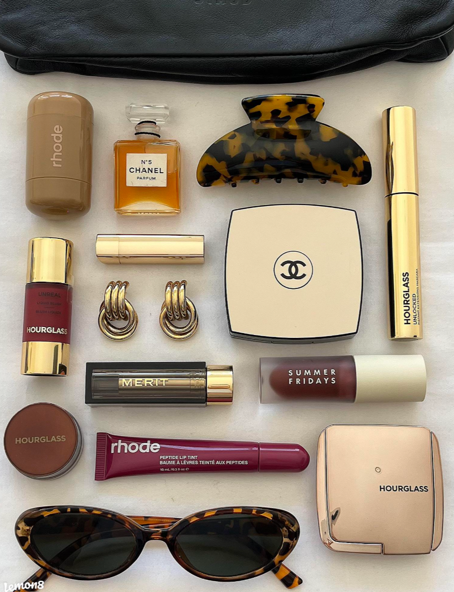 Top-down view of various fashion and beauty accessories, including a pair of sunglasses, lipstick, perfume bottles, hair clips, earrings, and cosmetic items, arranged on a white surface with a black bag partially visible in the background.