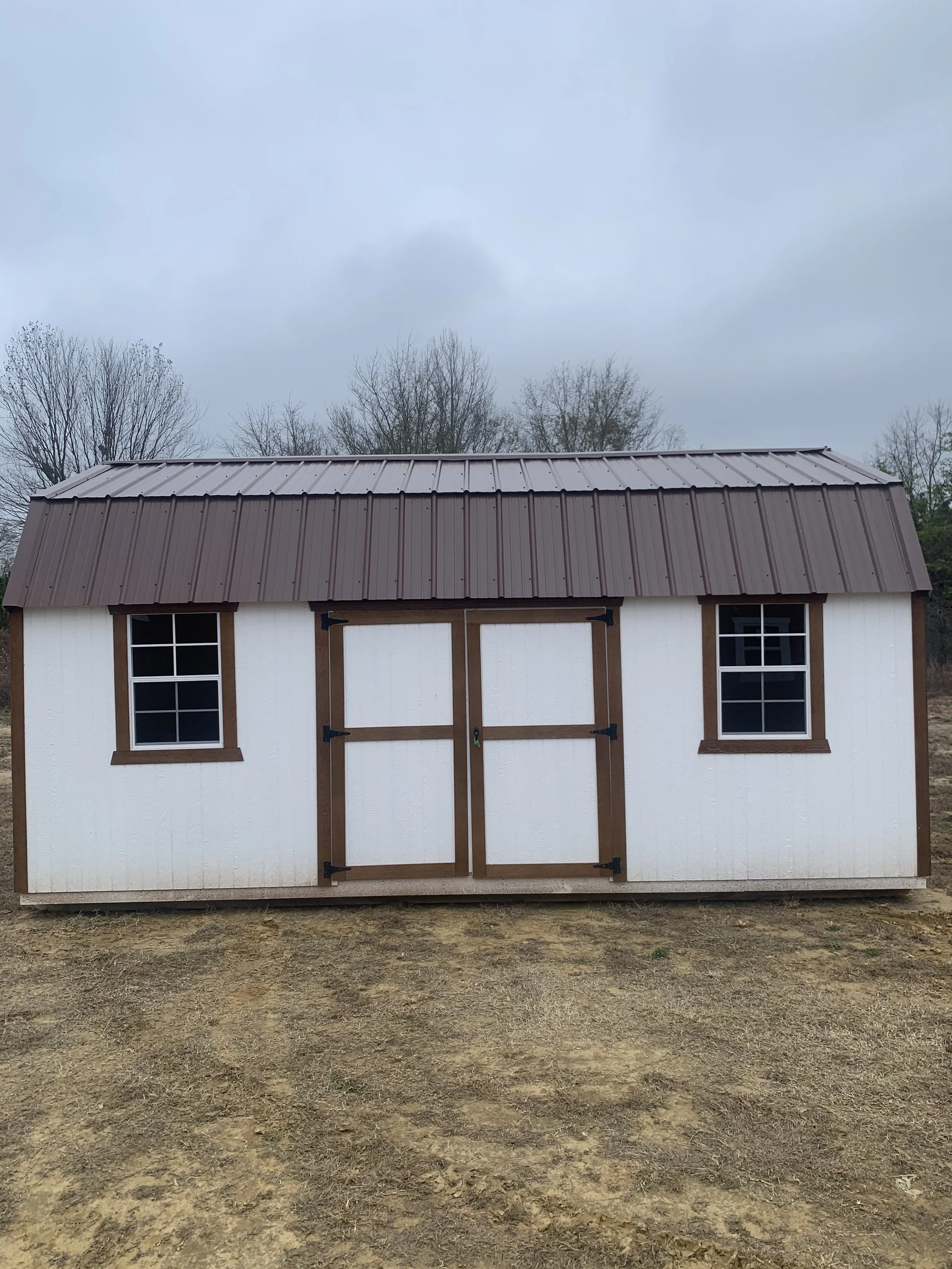 White outdoor shed with a brown metal roof, double doors, and two windows in a field.