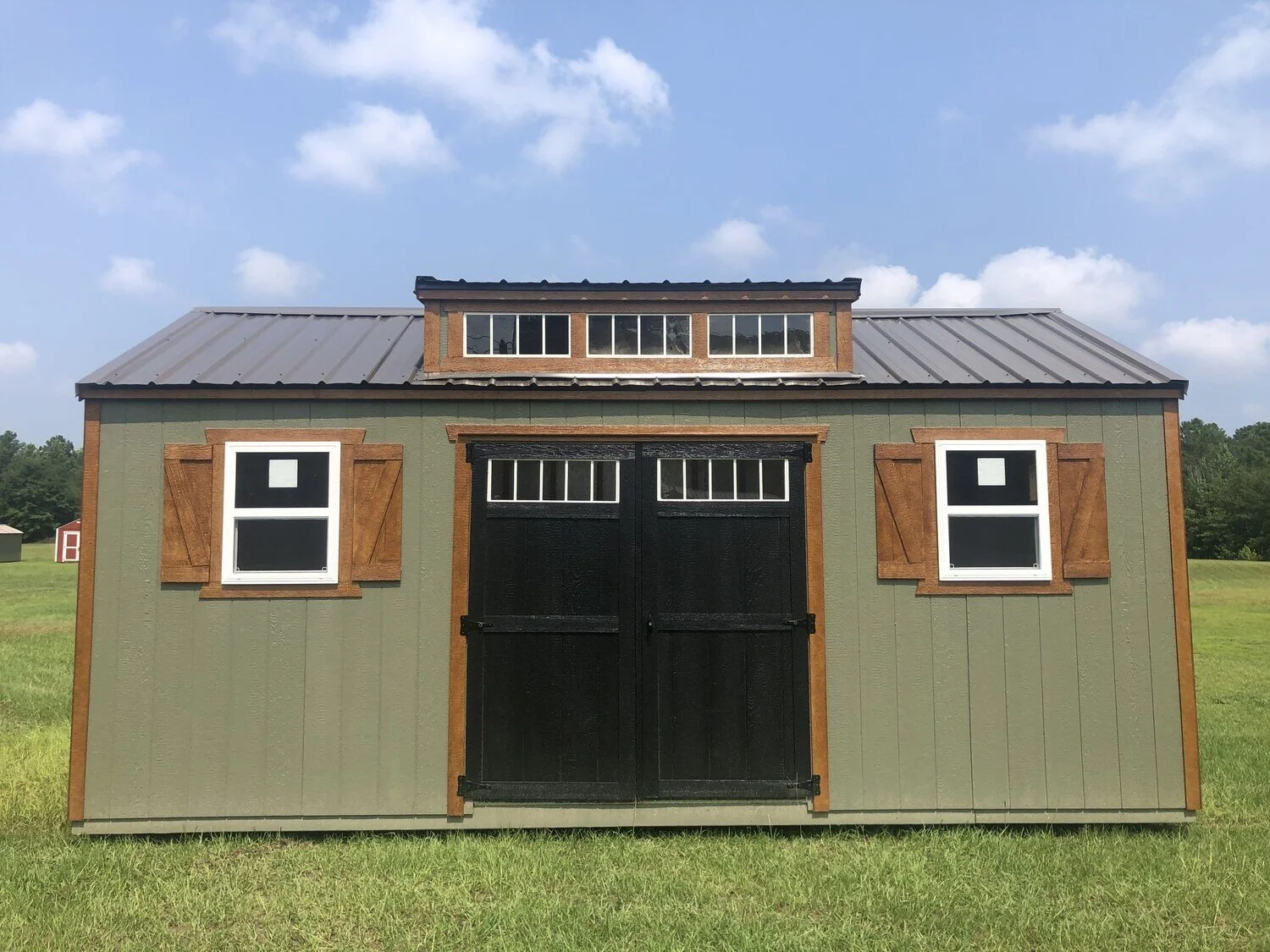 Green storage shed with brown accents and a black double door, featuring two windows on either side, in a grassy field.