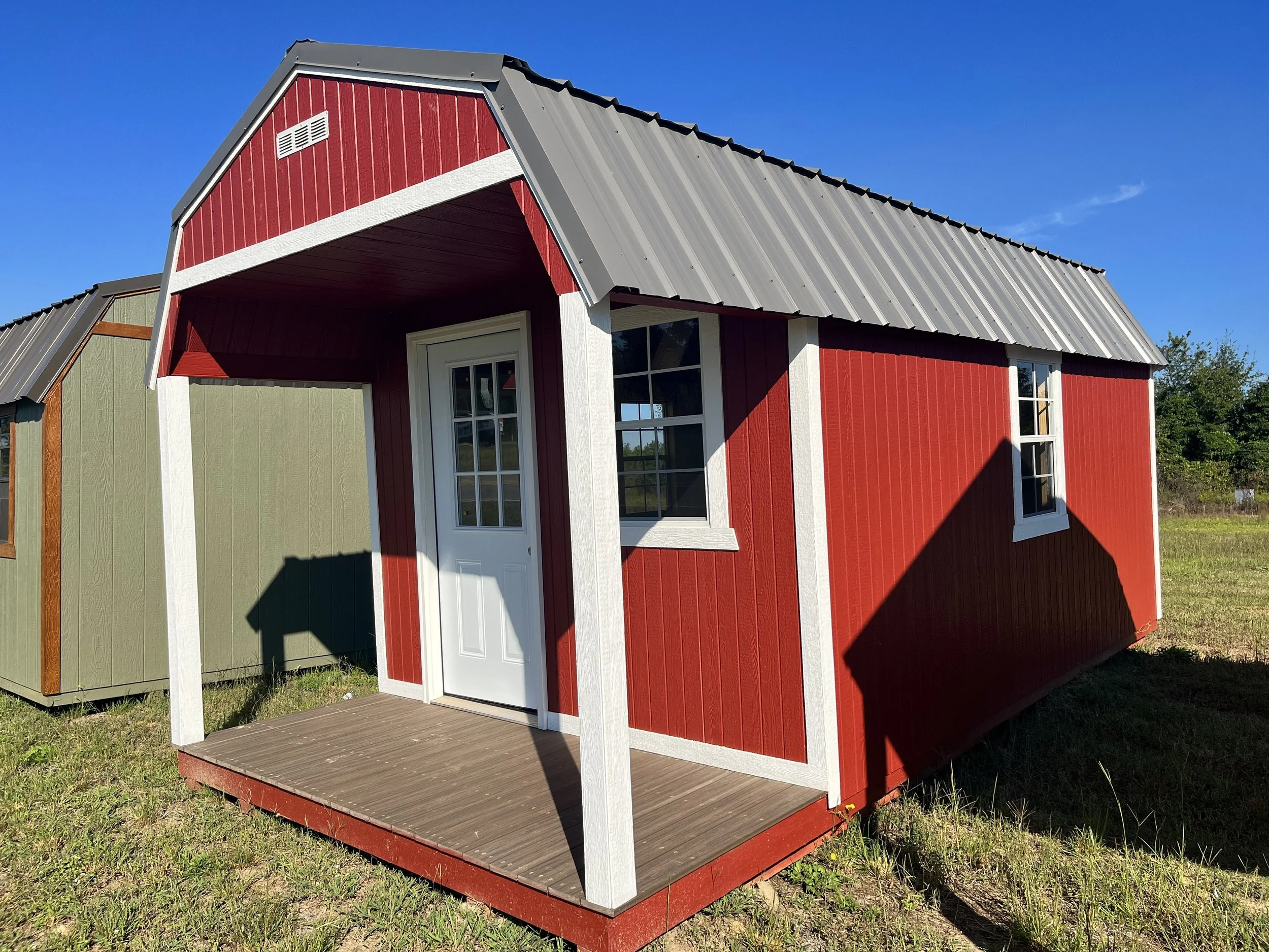 A small red and white shed with a gray metal roof and a front porch, set on grass under a clear blue sky.