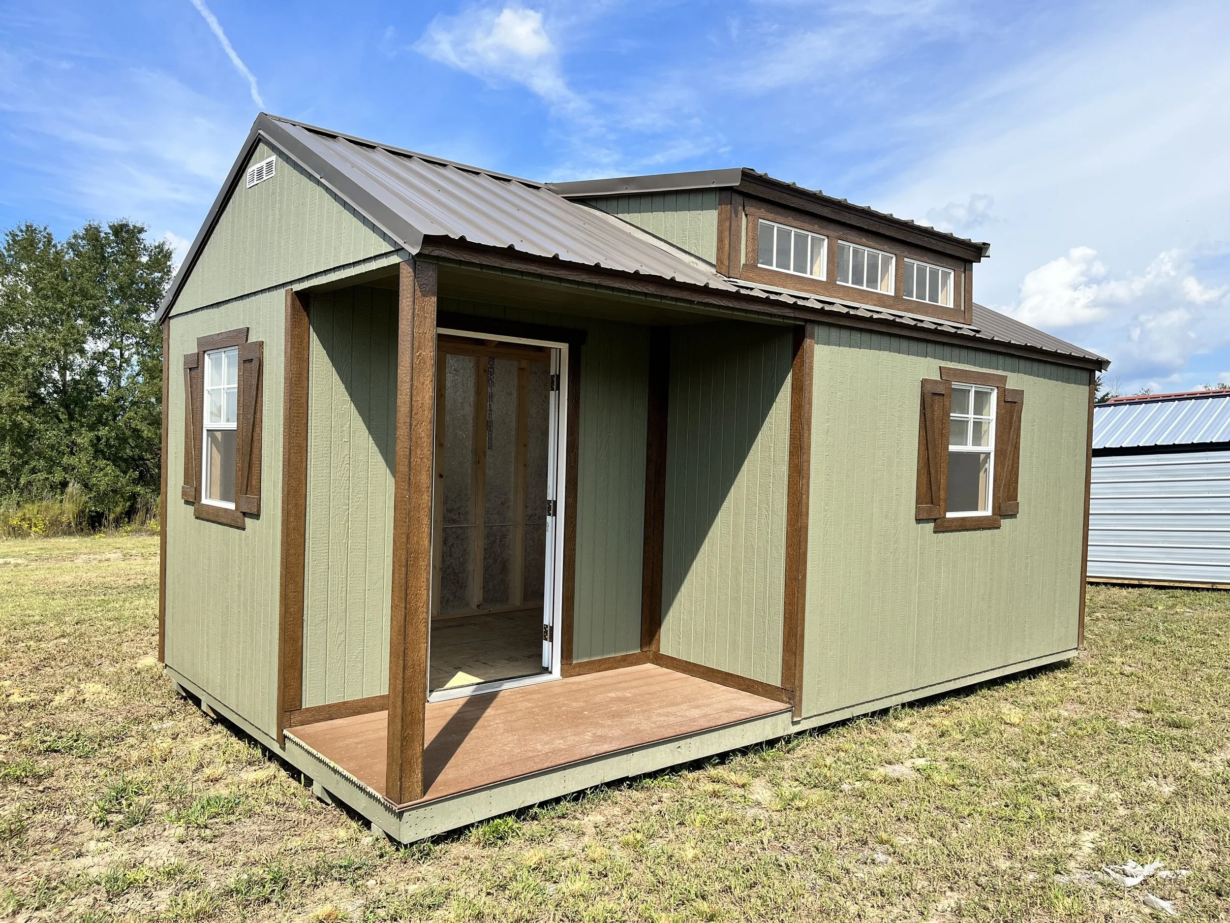 Small green shed with brown trim and windows, featuring a porch and metal roof, set on grassy land under a blue sky.