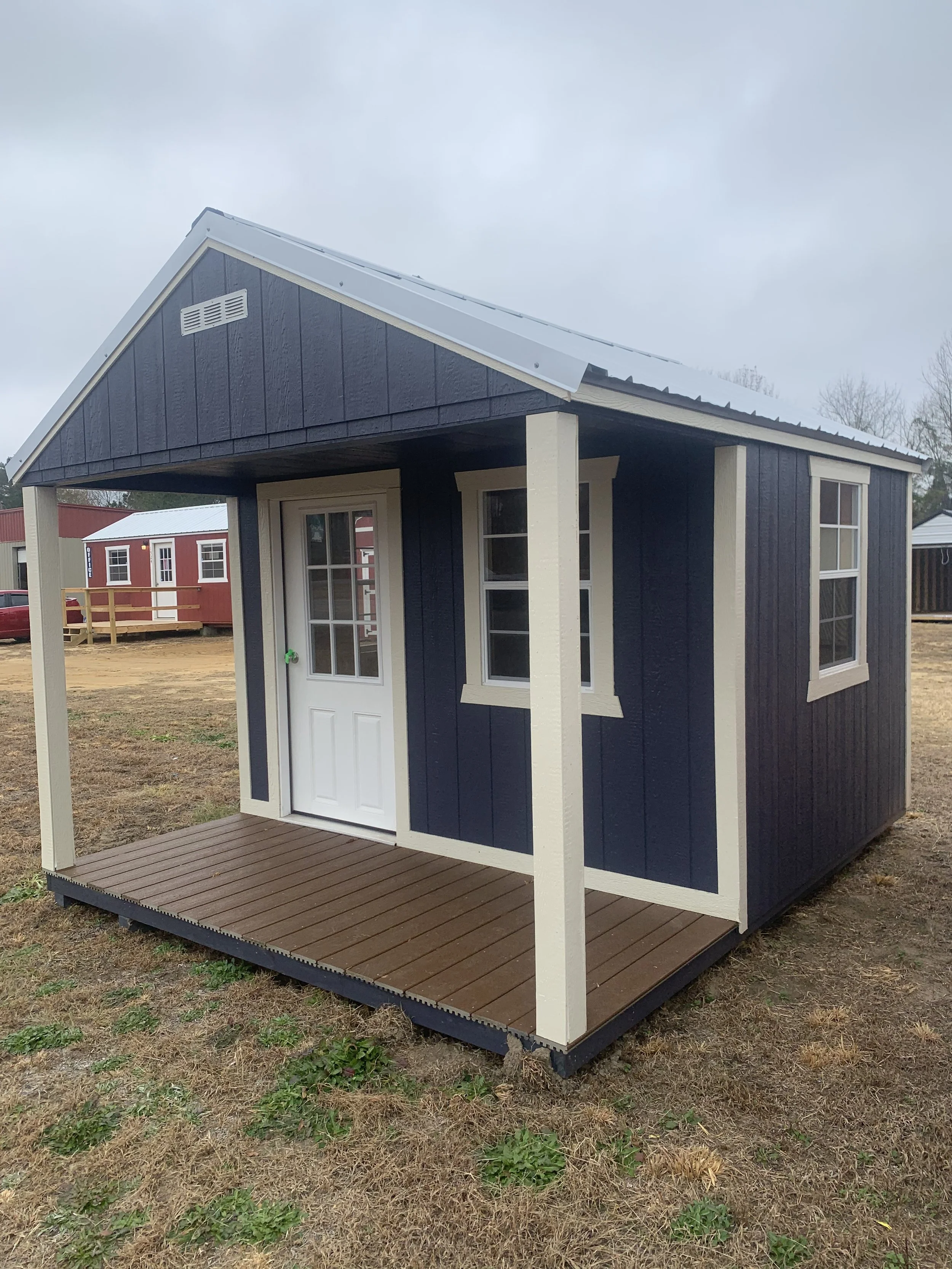 A small dark blue storage shed with a white door and white trim, featuring a pitched metal roof and a small covered porch on a grassy area.