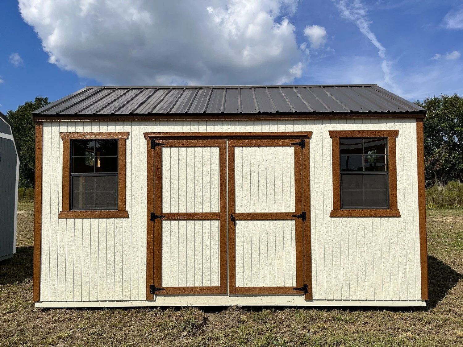 A small beige shed with a metal roof and brown wooden trim around the door and two windows, set on a grassy area under a partly cloudy sky.