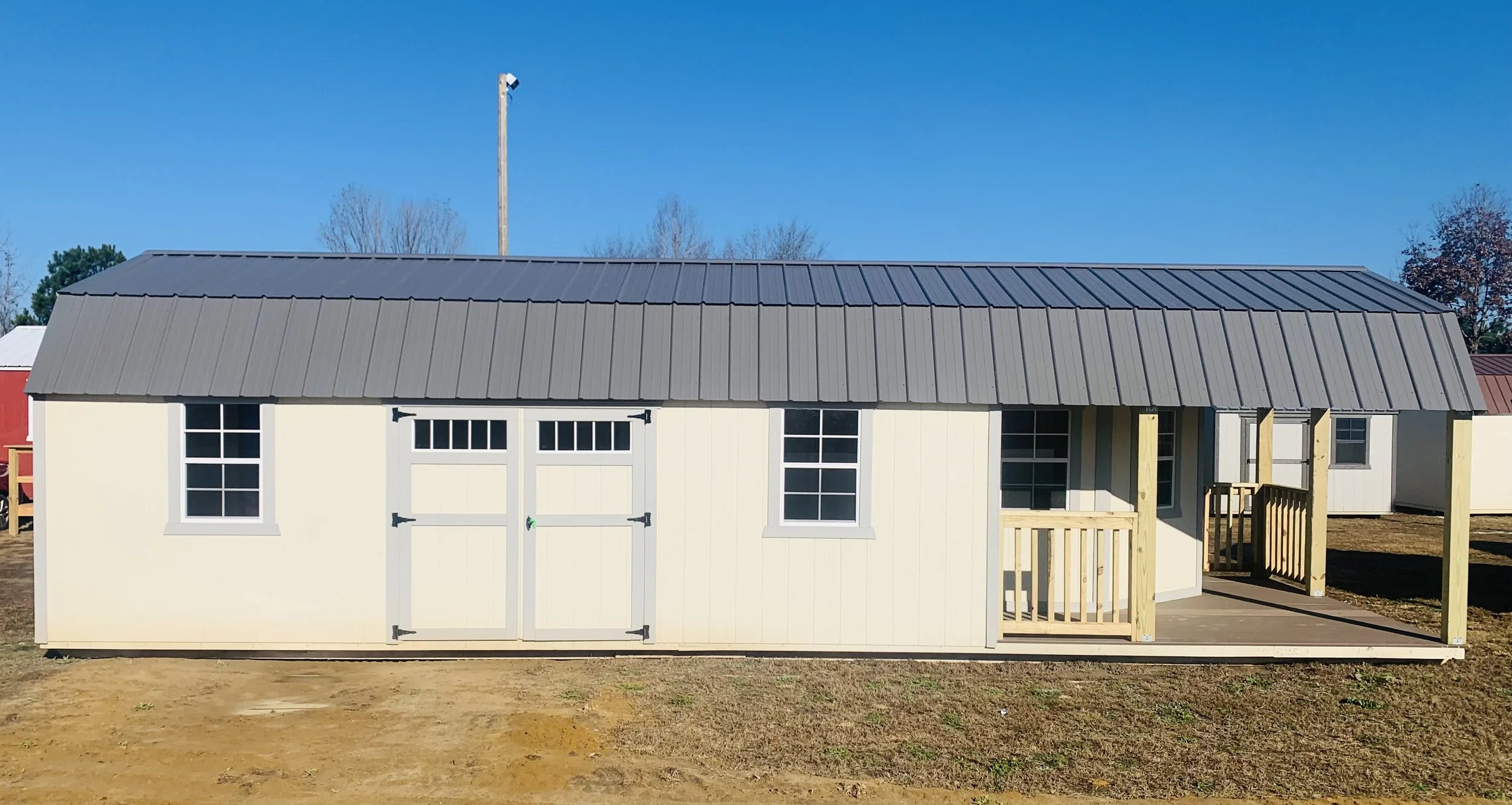 Small cream-colored shed with a gray metal roof and two windows on a wooden porch.