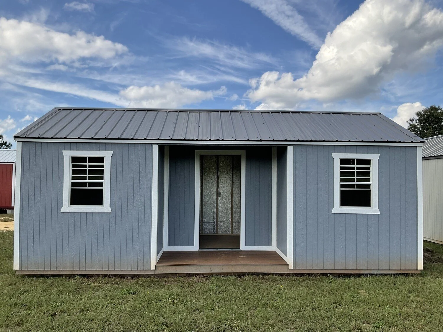 Small blue wooden cabin with a metal roof and two front windows on grass with a cloudy sky.