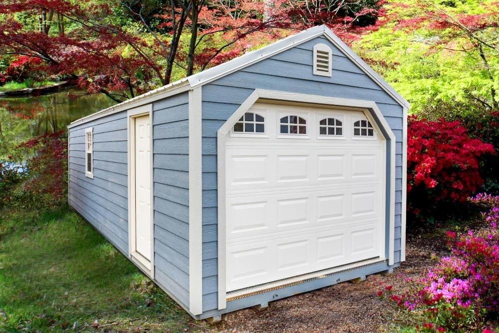 A small blue storage shed with a white garage door and side door, set in a garden with colorful flowers and trees.