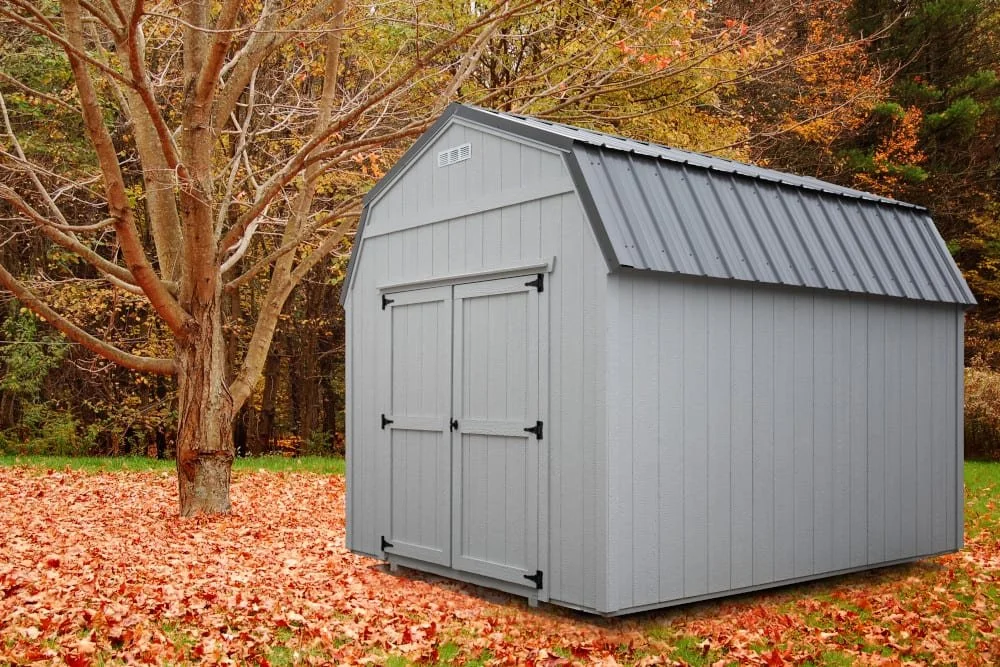 Gray storage shed with a sloped roof in a fall setting, surrounded by trees and fallen leaves.
