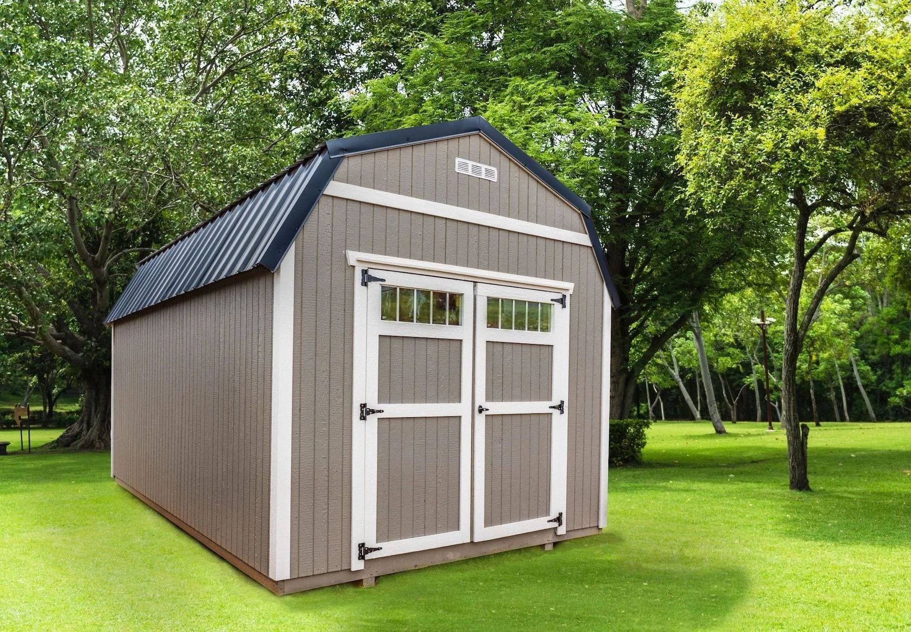 A brown storage shed with white trim and a black metal roof, situated on a green grassy lawn with trees in the background.
