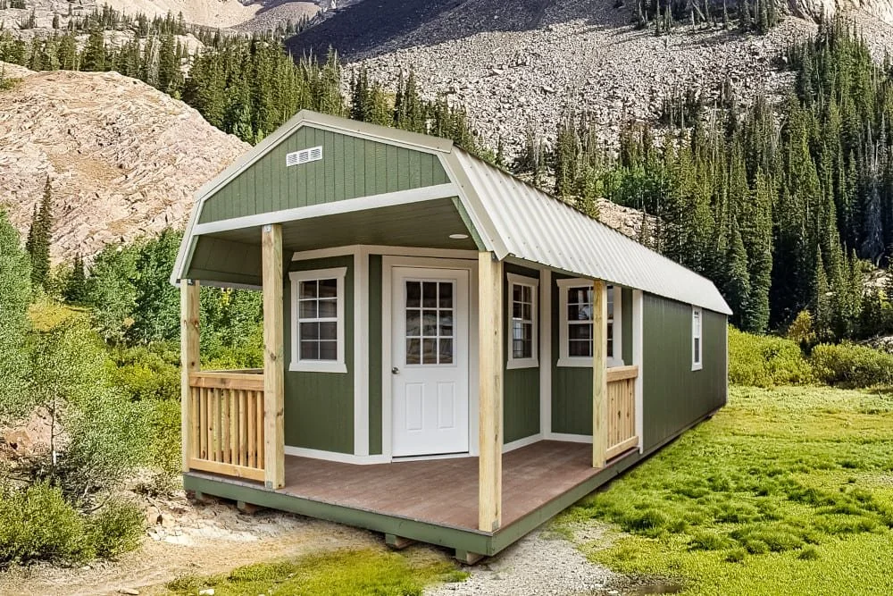 A small, green wooden cabin with a white roof and porch set in a mountainous landscape with pine trees.
