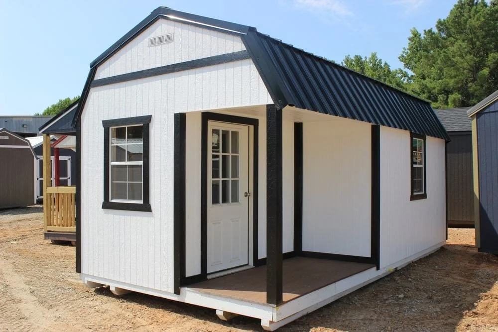 Portable white cabin with a black metal roof, featuring a door and windows, displayed outdoors.