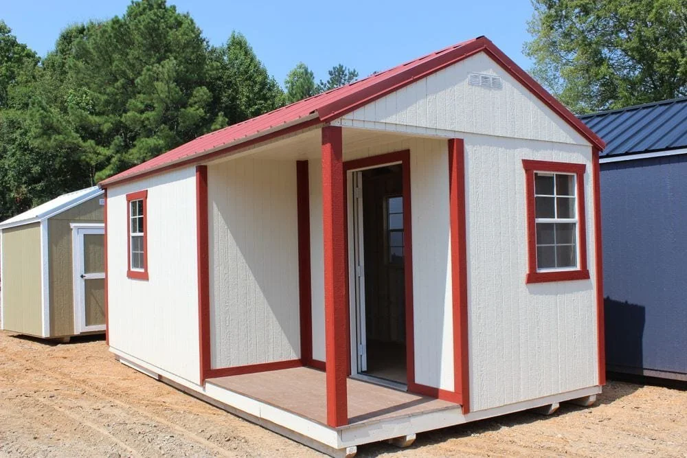 Small portable building with a beige exterior and red trim, featuring two windows and an open door, set on a gravel surface with trees in the background.