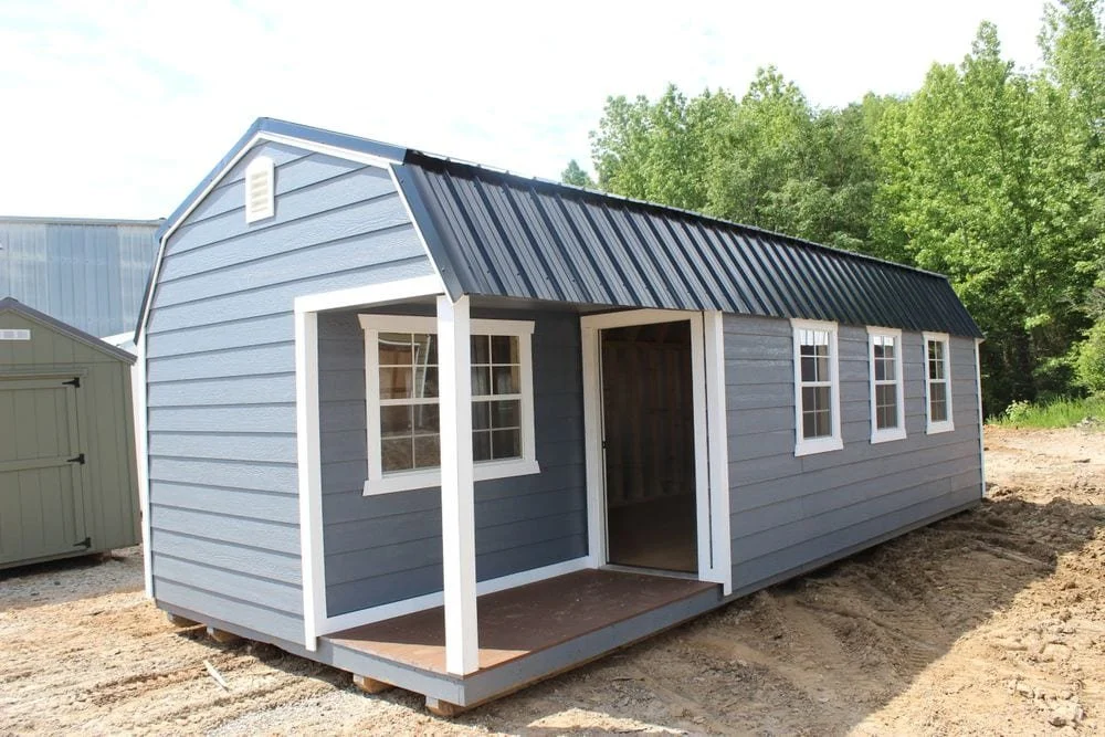 Blue shed with white trim and four windows, featuring a black metal roof, situated outdoors on a dirt area with trees in the background.