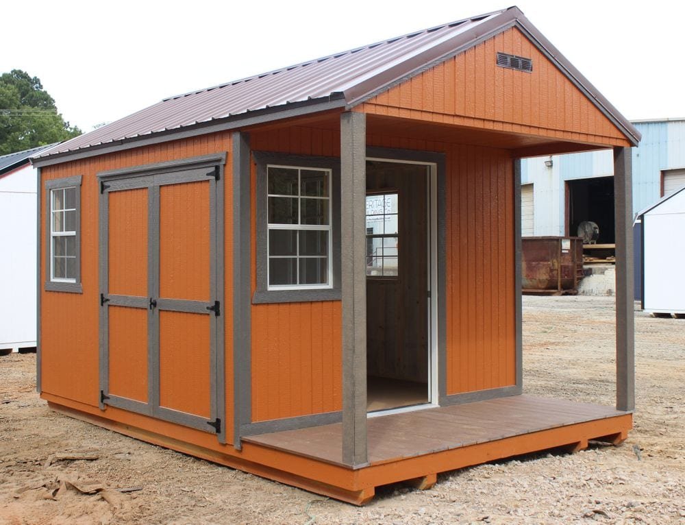 Wooden storage shed with orange and brown siding, featuring double doors and windows, set on a dirt lot with industrial buildings in the background.