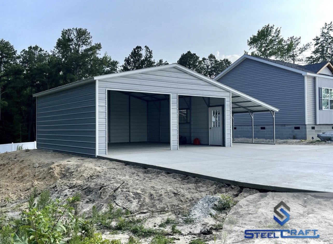 Steel building garage with open doors on a concrete driveway next to a house, surrounded by trees.