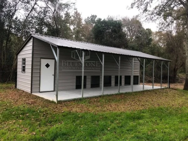 Gray metal building with a white door and open garage-style space, set on sandy ground with trees in the background under a partly cloudy sky.