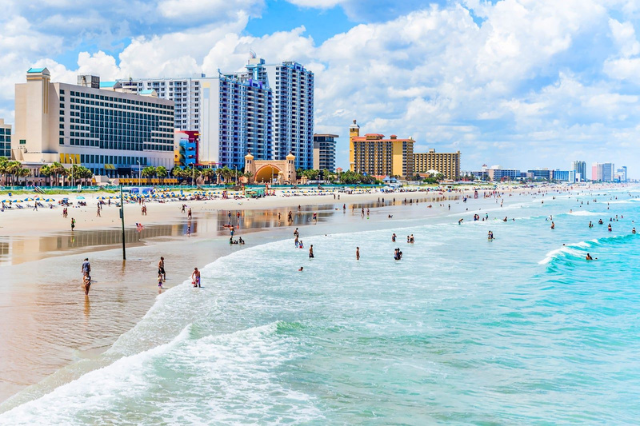Beach scene with people swimming and walking along the shoreline, with high-rise hotels and buildings in the background, under a partly cloudy sky.