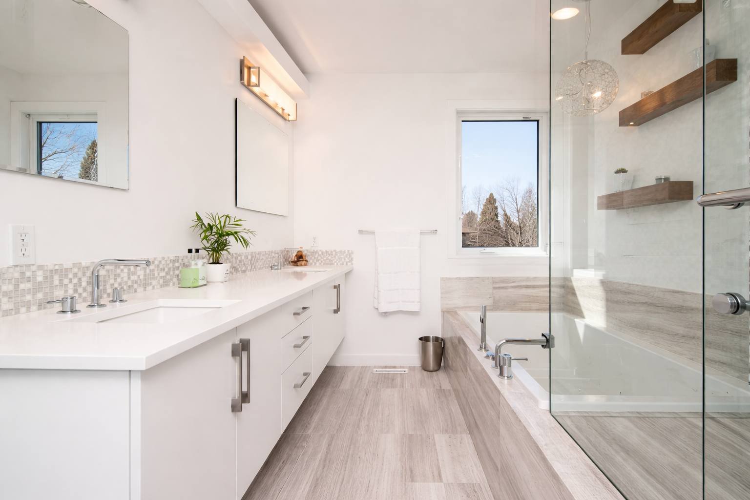 Modern bathroom with white cabinetry and beige tiled floor. Features a bathtub with a glass enclosure, a window showing trees outside, and a white countertop with a sink, mirror, and decorative items.