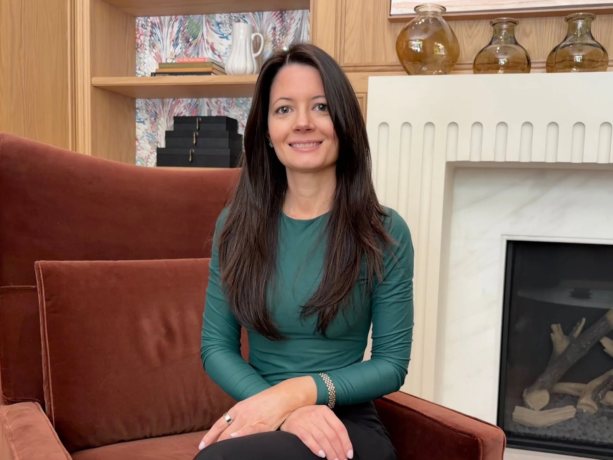 Nicole Cleary, health coach and founder of Nourished by Nicole — A woman with long dark hair wearing a teal long-sleeve top sitting on a reddish-brown sofa in a warmly decorated room.