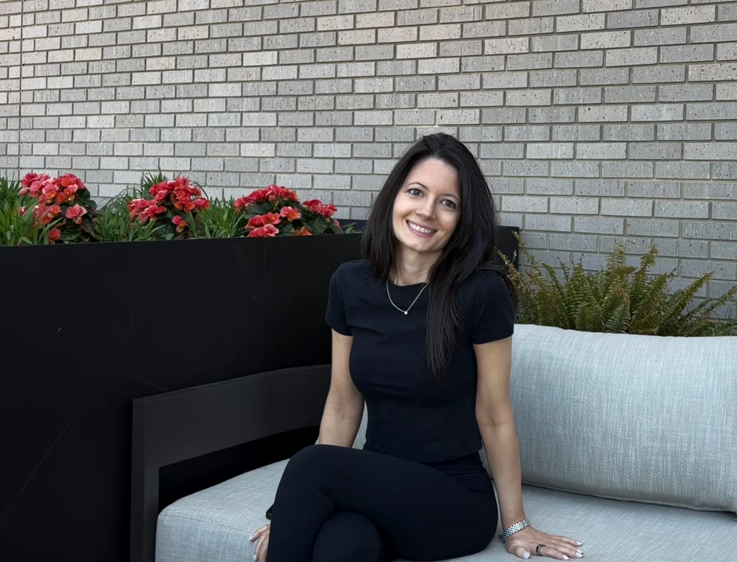 Nicole Cleary, IIN certified health coach and lifestyle architect — A woman with long dark hair smiling while sitting on a light gray outdoor sofa with pink and red flowers behind her.