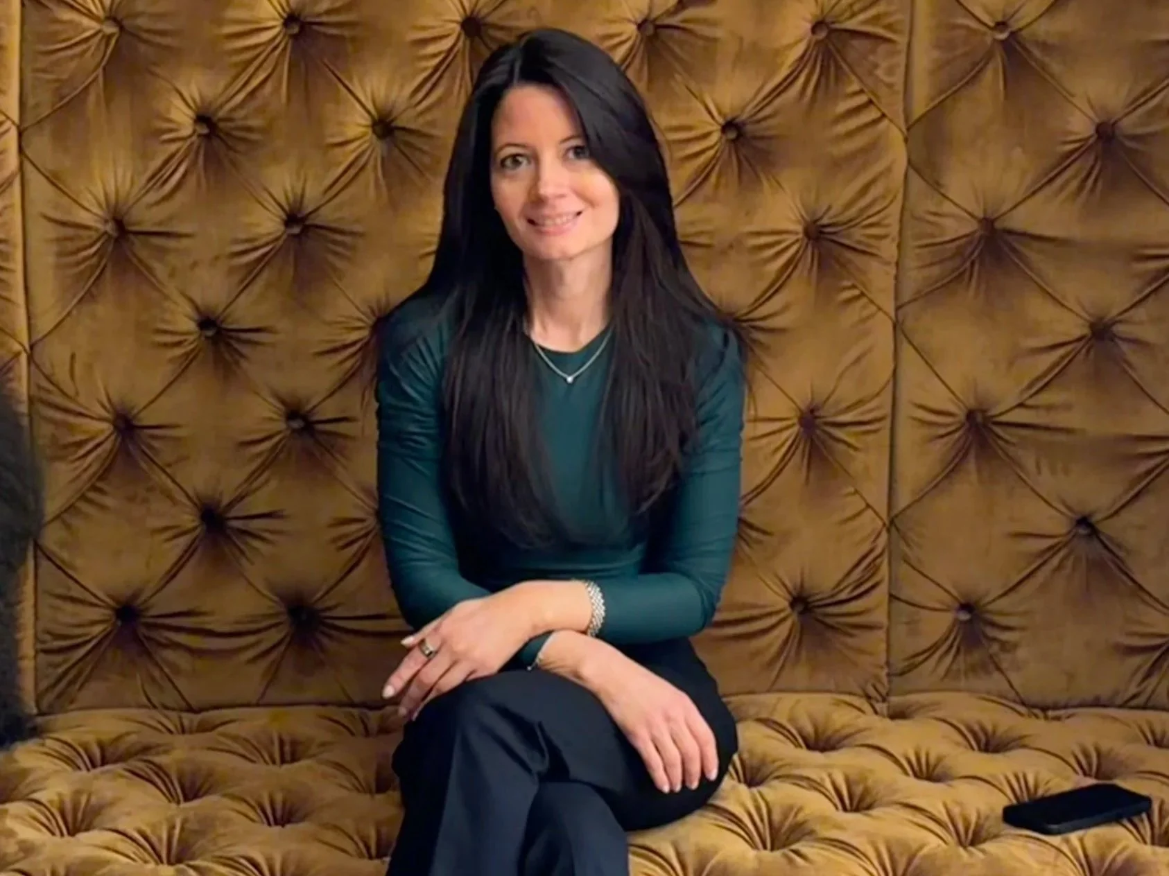 Nicole Cleary, health coach and founder of Nourished by Nicole​​​​​​​​​​​​​​​​ - A woman with long dark hair wearing a teal long-sleeve top and black pants seated and smiling at the camera on a sofa.