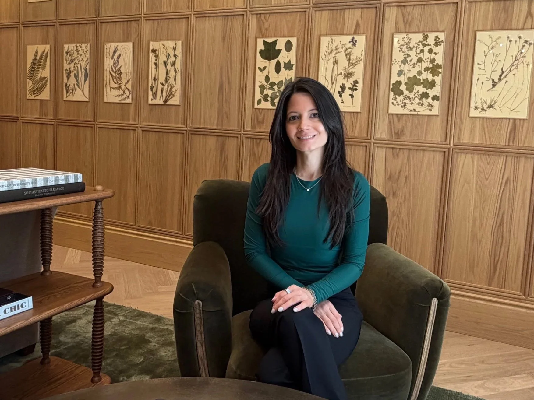 A woman with long dark hair wearing a teal top seated and smiling in a green armchair in a room with wood-paneled walls — Nicole Cleary, habit strategist and wellness coach, Nourished by Nicole