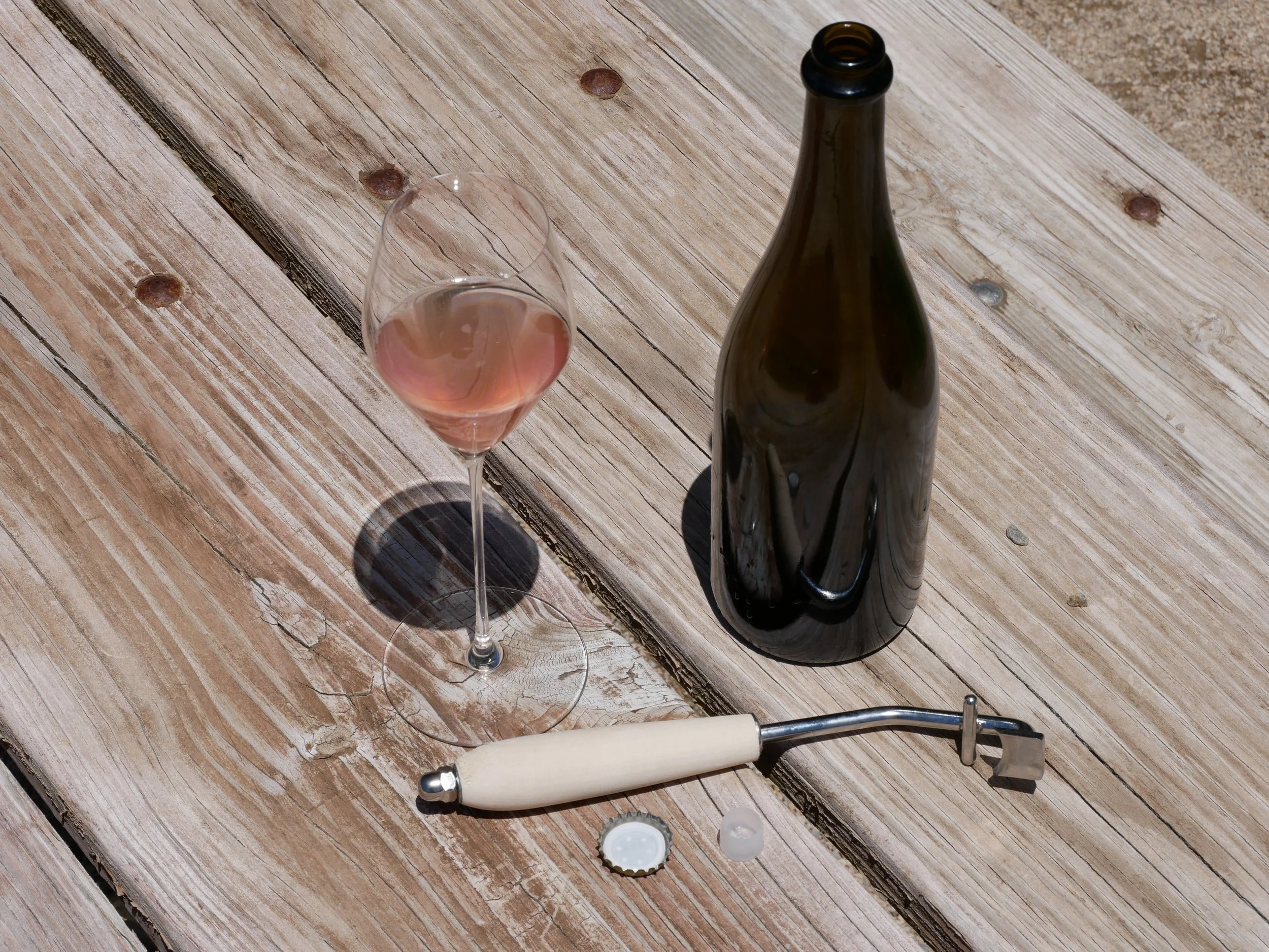 A glass of AERILYN 2025 sparkling rosé beside the opened bottle, disgorgement key, crown cap, and bidule after the first trial disgorgement, July 2026.