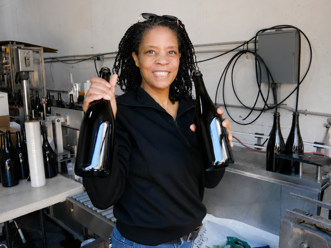Tammee Thompson, founder of AERILYN Wine Co., holding two bottles of the 2025 vintage on tirage bottling day at the production facility in Orcutt, CA.