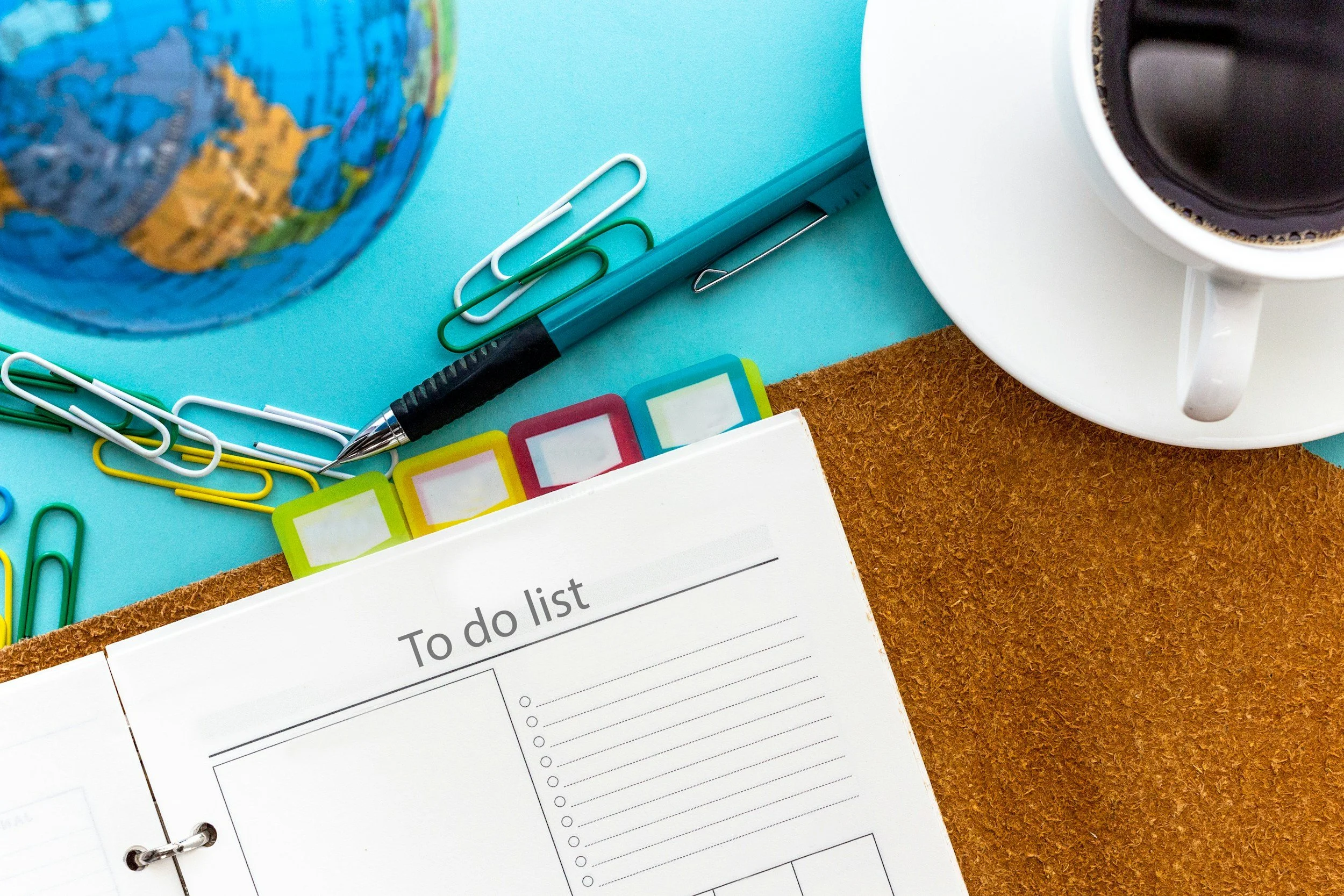 A colorful desk with a globe, paper clips, a pen, sticky notes, a to-do list notebook, a cup of coffee, and a cork mat.