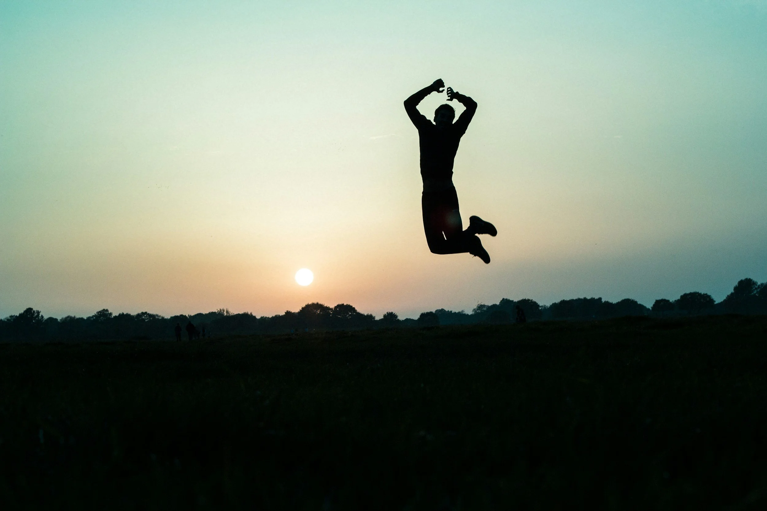 Silhouette of a person jumping in the air during sunset with the sun low on the horizon and trees in the background.