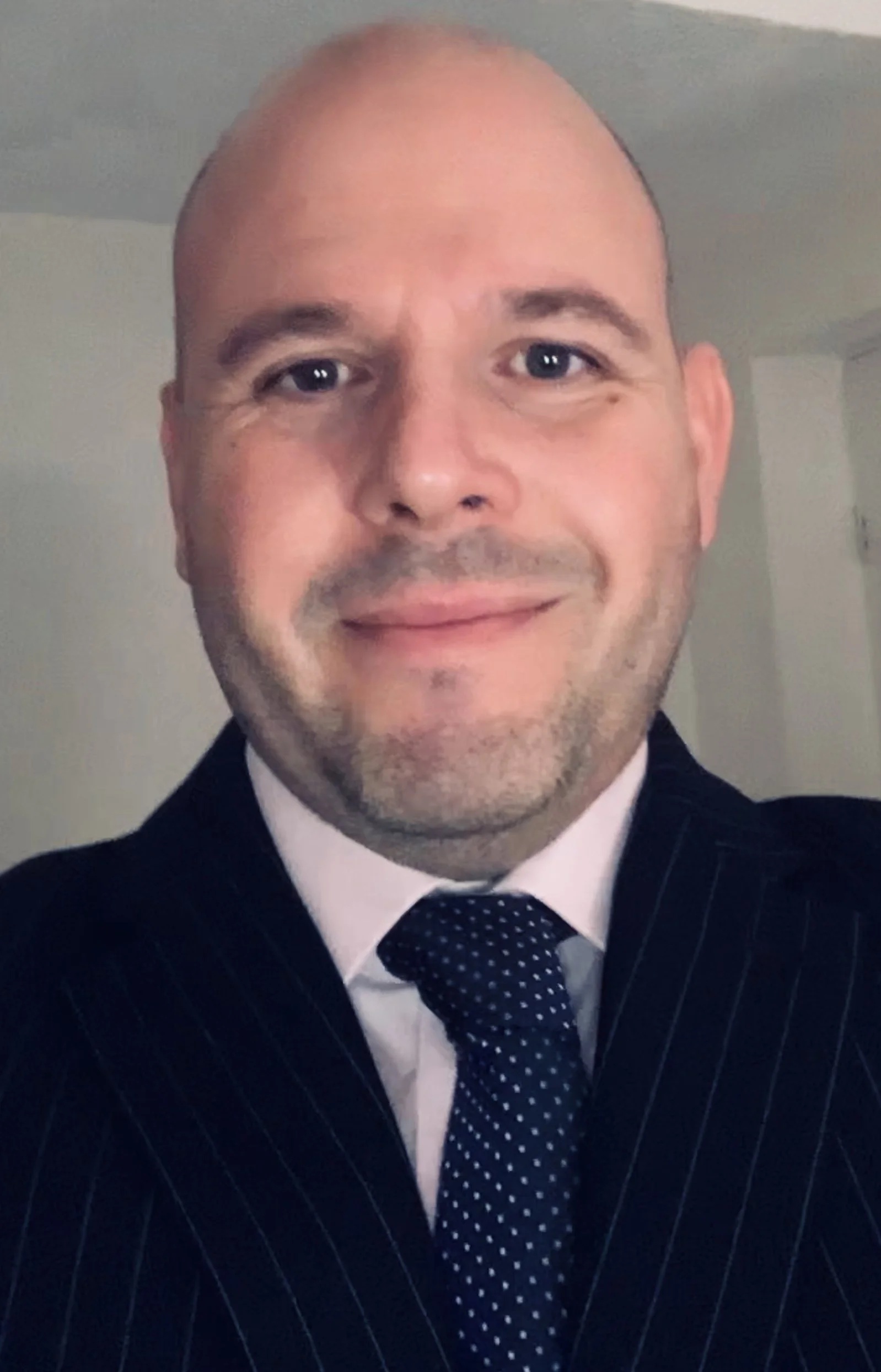 A smiling, bald man wearing a dark pinstripe suit, white shirt, and a navy blue tie with small white polka dots.