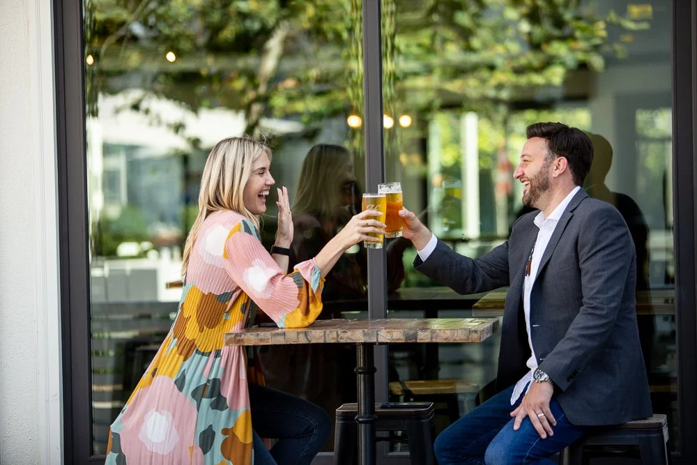 A man and woman sitting at a table outside a cafe, clinking glasses of beer and smiling.