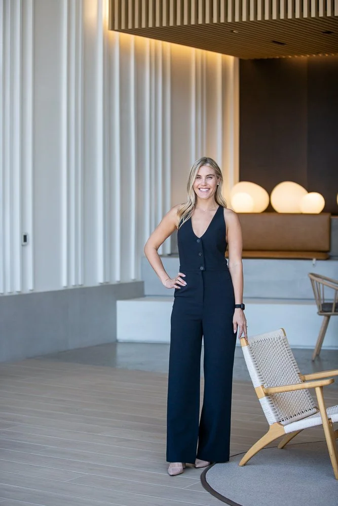 A woman with blonde hair smiling in a navy jumpsuit standing indoors near a woven chair, with modern decor and soft lighting in the background.