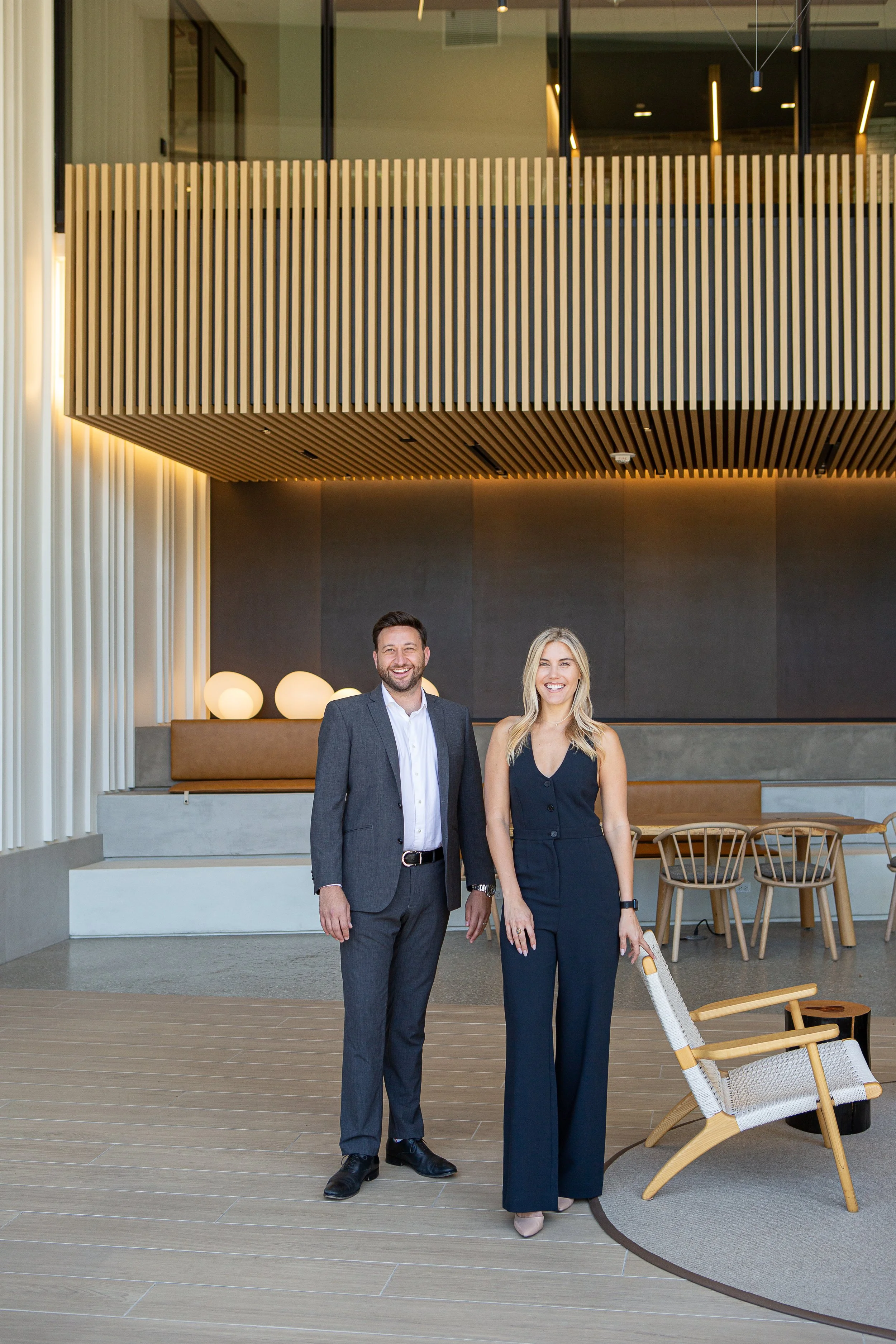 A man and woman in formal attire smiling and standing inside a modern building with wood accents, contemporary lighting, and stylish furniture.