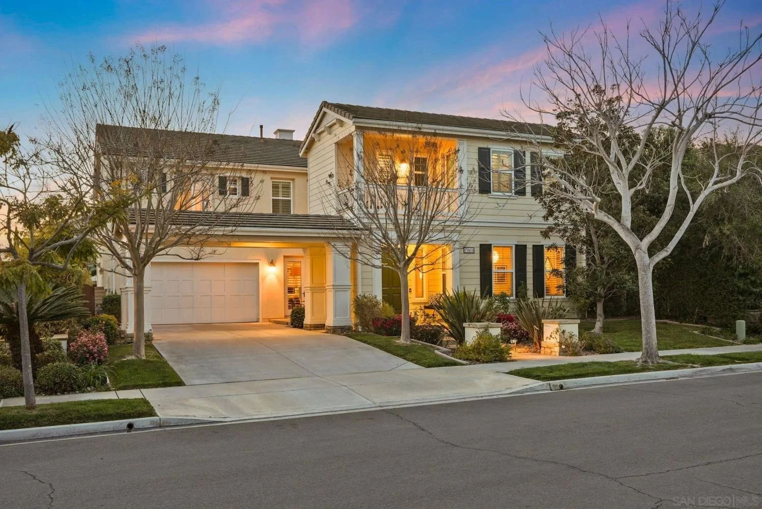 A two-story house with a well-maintained front yard, trees, and shrubs, illuminated with warm interior and exterior lighting during dusk.