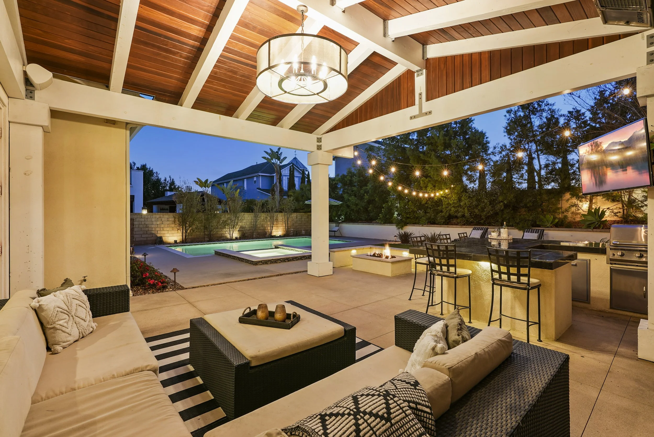 Outdoor living space at dusk with patio furniture, string lights, a firepit, swimming pool, and a television mounted on the wall.