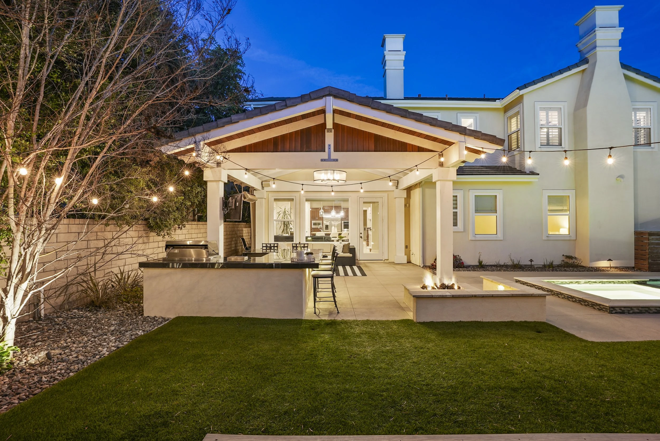 Backyard patio area illuminated with string lights, featuring a built-in outdoor kitchen with a bar counter, and a hot tub in the right corner. The background shows a large white house with multiple windows and a blue evening sky.