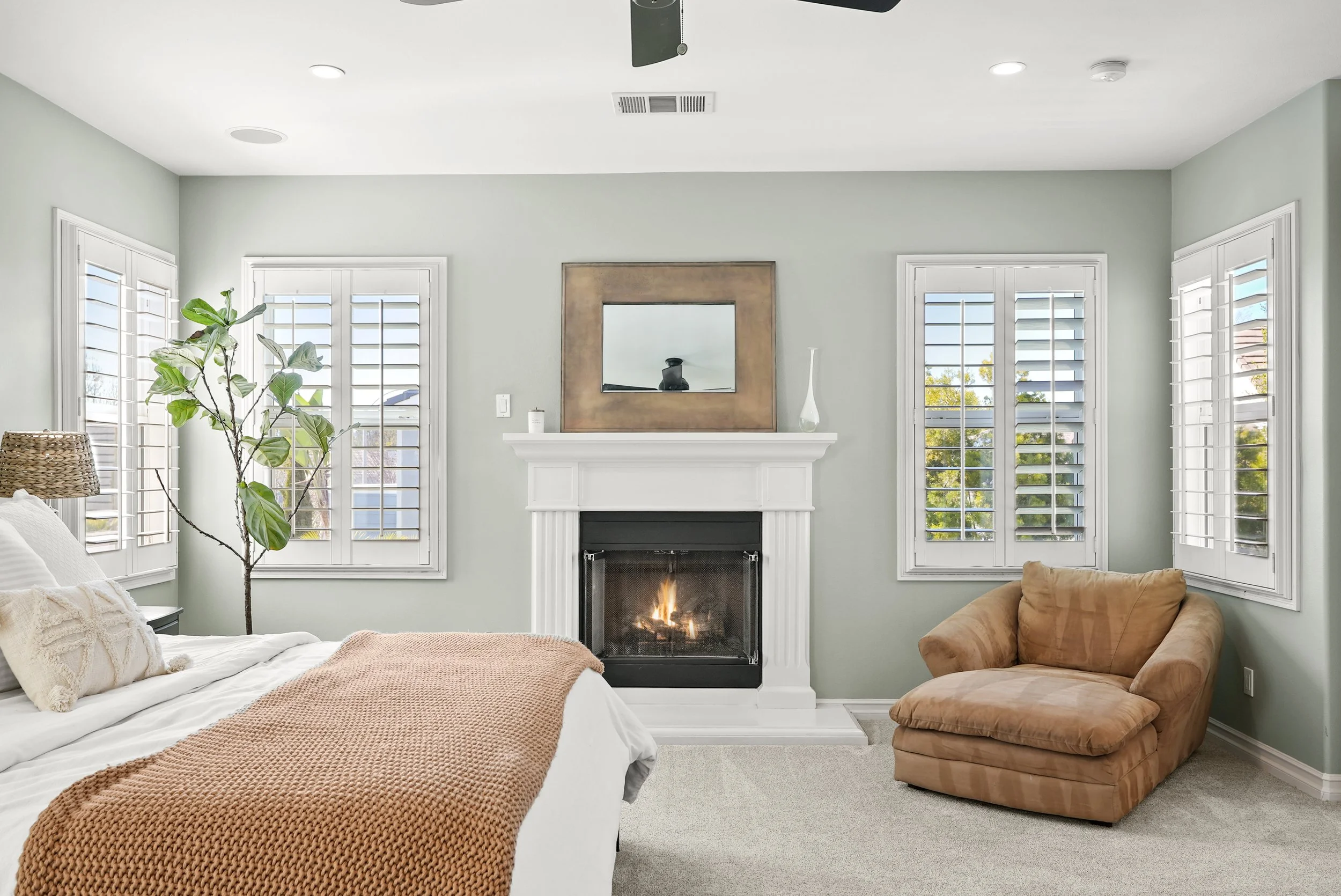 Living room with a fireplace, beige armchair, white sofa, large window with white shutters, a large potted plant, and a mirror on the wall above the fireplace