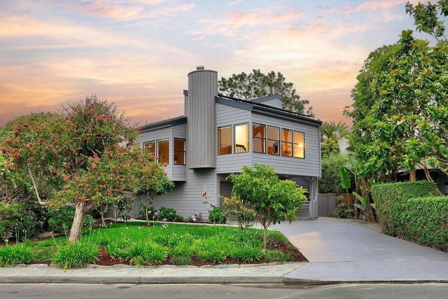 A modern two-story house painted in gray with large windows, surrounded by lush green trees and plants, at sunset.