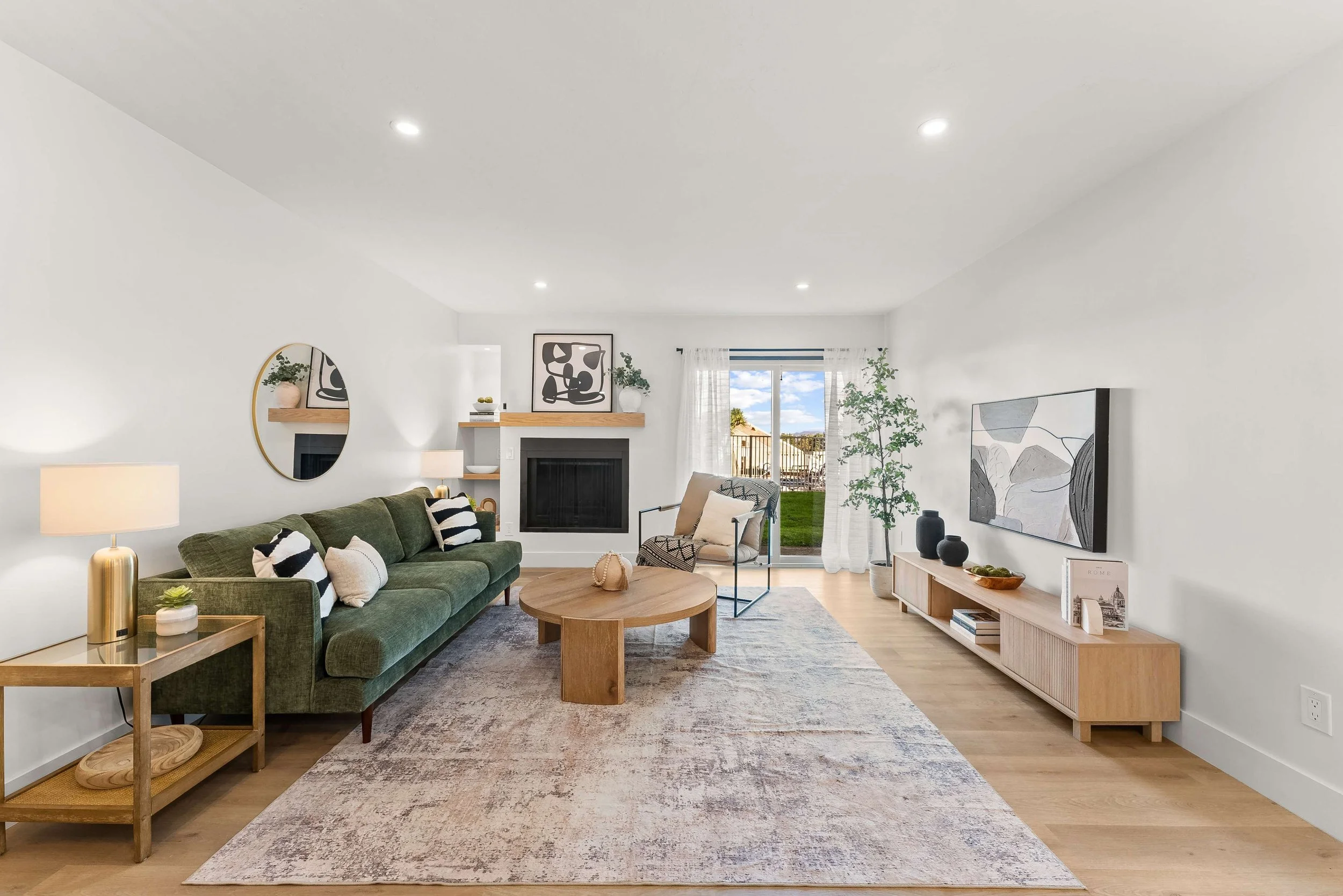 A living room with a green sofa, a rustic wooden coffee table, and a beige armchair near sliding glass doors leading outside. Decor includes black and white pillows, a wall-mounted mirror, framed art, and potted plants, with a modern minimalist style and natural light.