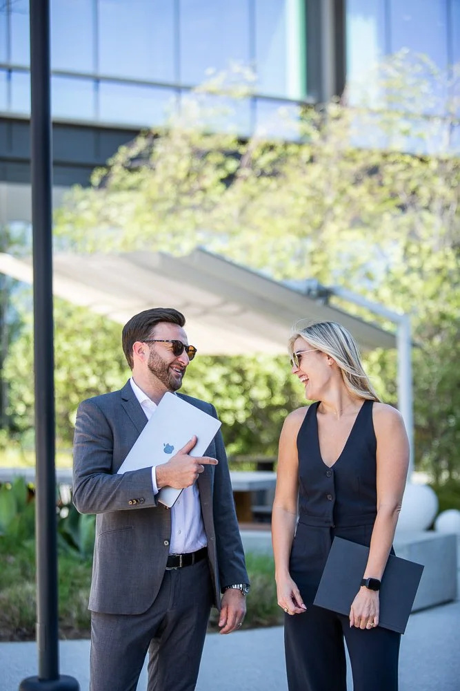 Businessman and businesswoman talking and smiling outside, holding laptops, with modern office building and trees in the background.