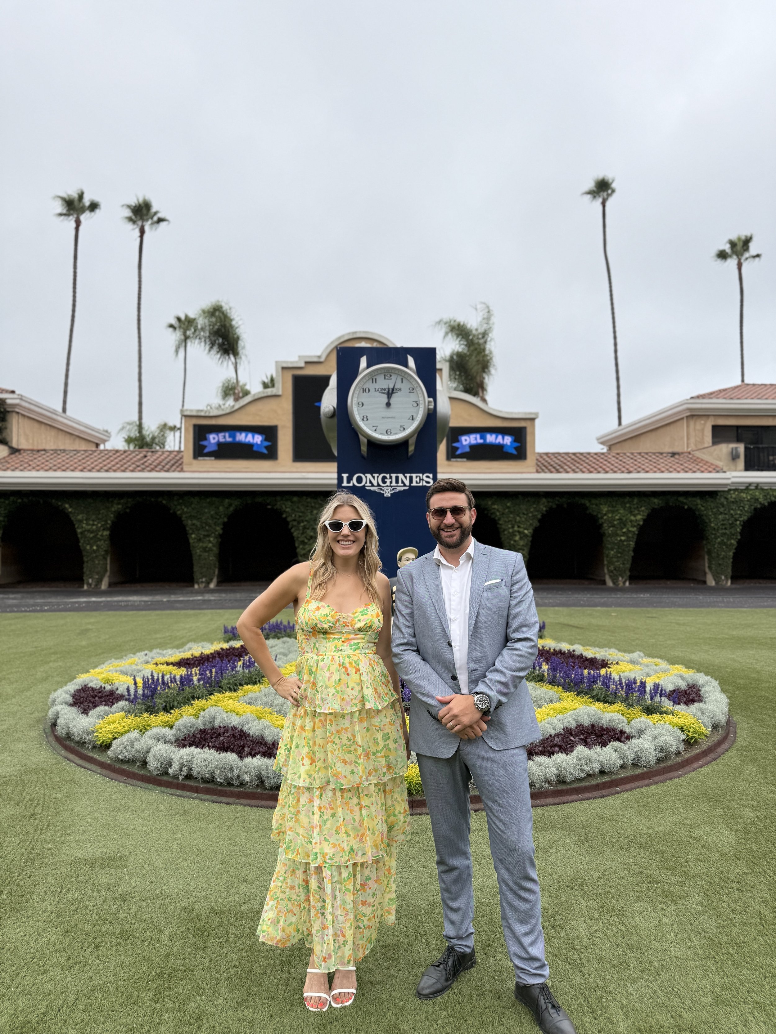 A woman in a yellow floral dress and sunglasses standing next to a man in a gray suit and black shoes, both smiling, in front of a flower bed and a large clock display at Del Mar racetrack, with tall palm trees and a cloudy sky in the background.