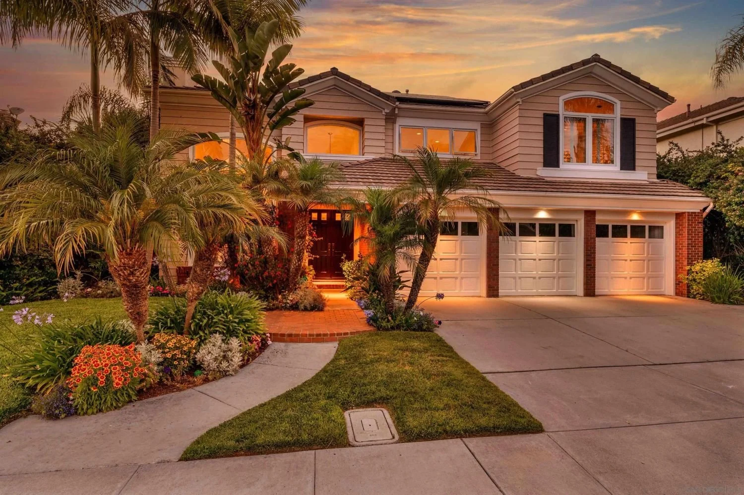 A two-story suburban house during sunset with palm trees and landscaped front yard featuring colorful flowers and lush grass, with a concrete driveway leading to a three-car garage.