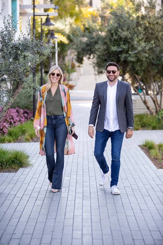 A woman and man walking together outdoors on a paved path, smiling and wearing sunglasses. The woman is in a colorful shawl and jeans, while the man is in a gray blazer, white shirt, and jeans. There are trees and bushes on either side of the path.