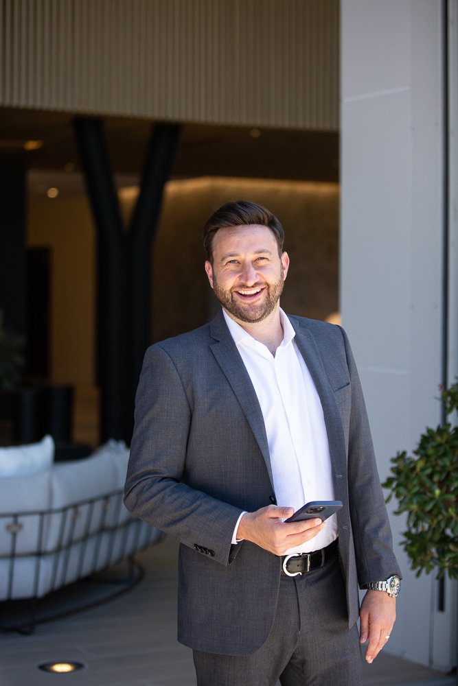 A smiling man in a gray suit holding a smartphone, standing in a modern indoor setting with a sofa and vertical black beams in the background.