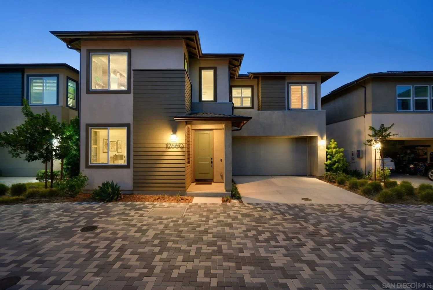 Modern two-story house with a front porch, illuminated exterior lighting, grey garage door, and landscaped front yard with small trees and plants at dusk.