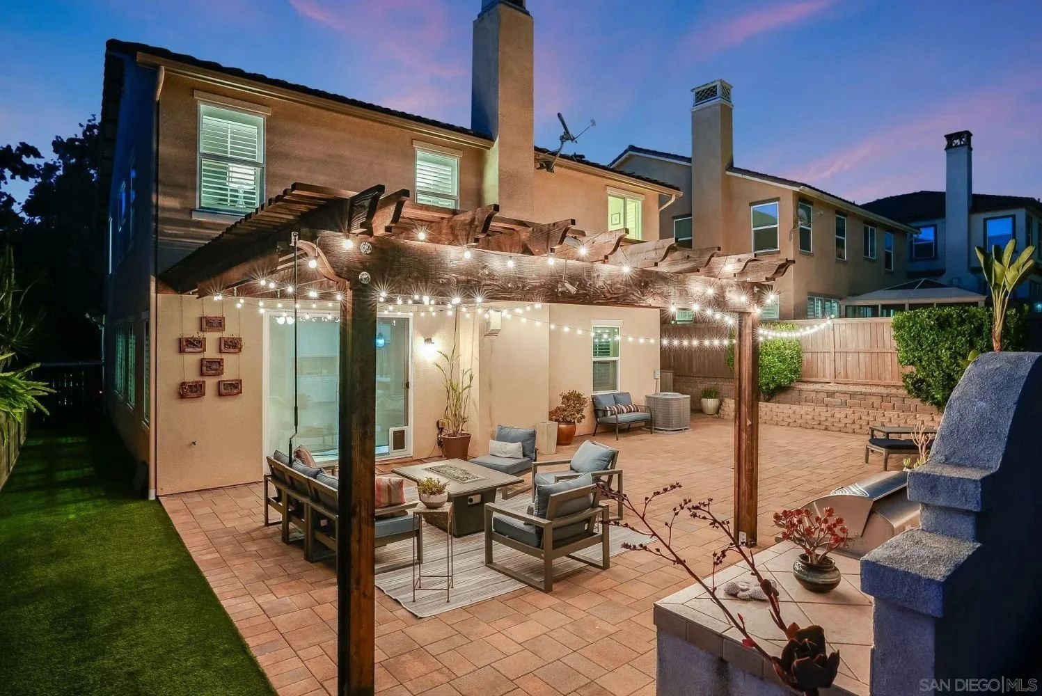 A home's backyard patio at dusk with string lights overhead, outdoor sofa and chairs, potted plants, and a concrete firepit, surrounded by a brick wall and neighboring houses.