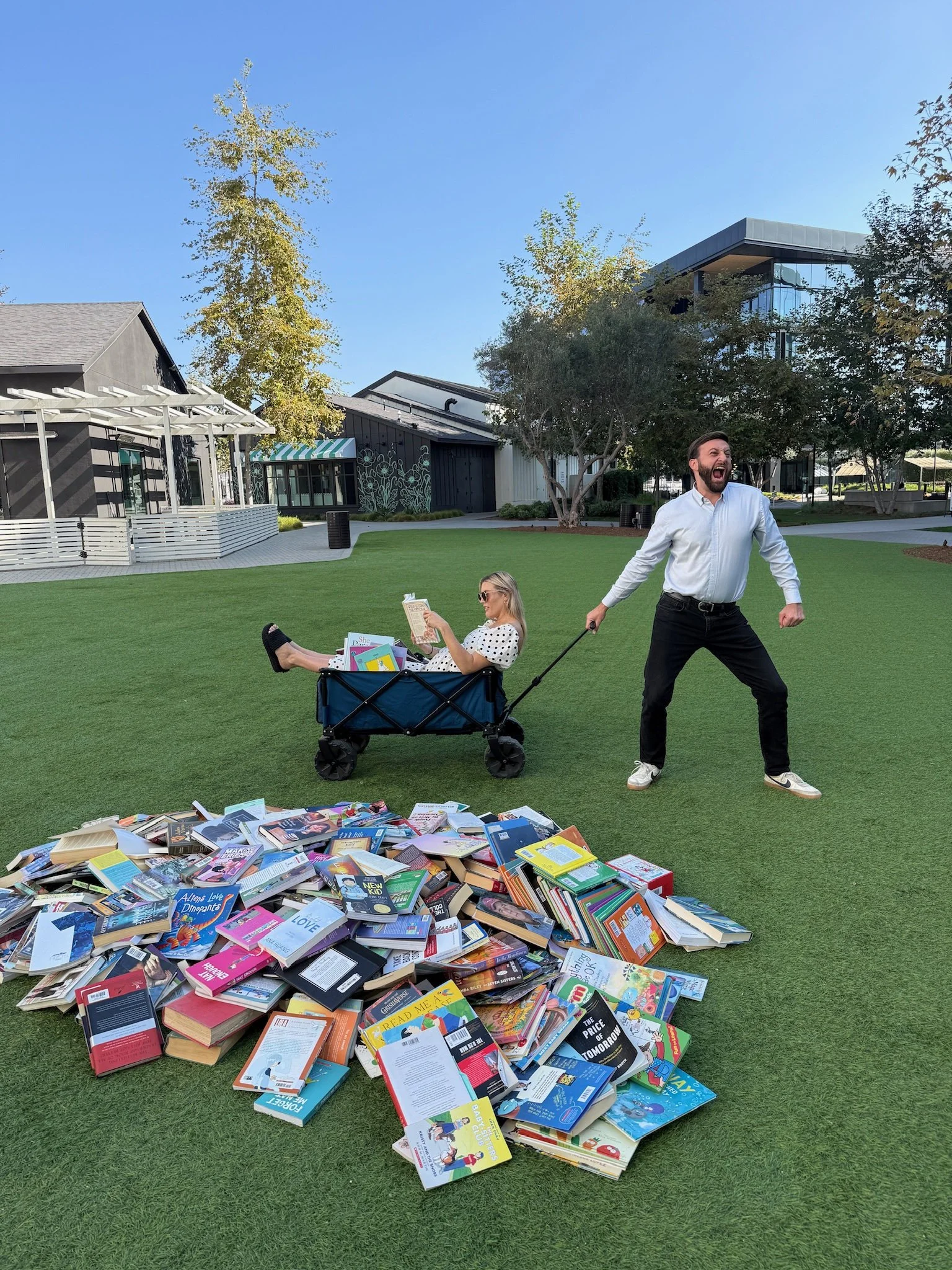 A man pulling a woman in a wagon filled with books on a grassy park area while the woman reads a book. There is a large pile of books on the ground in front of them. The background features trees and modern buildings.